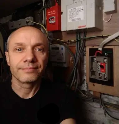 A professional commercial electrician in Plymouth standing next to a utility board installation featuring a mains switch, fire alarm control unit, and complex electrical wiring during a building inspection.